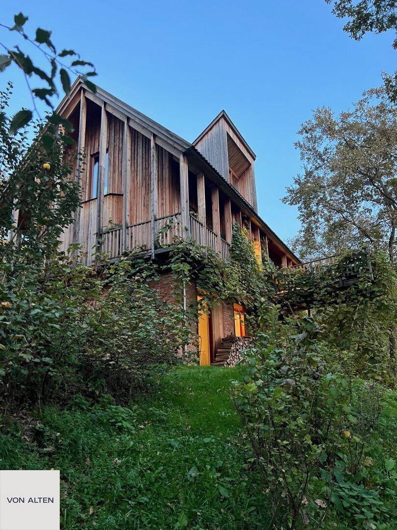Das Holzhaus in der Dämmerung, umgeben von üppiger Vegetation und sanften Hügeln.
