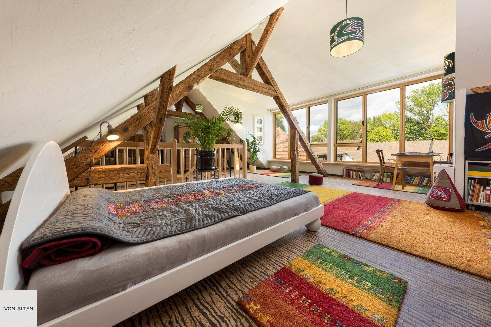 Gemütliches Schlafzimmer im Dachgeschoss mit Holzbalken, großen Fenstern und farbenfrohen Teppichen.