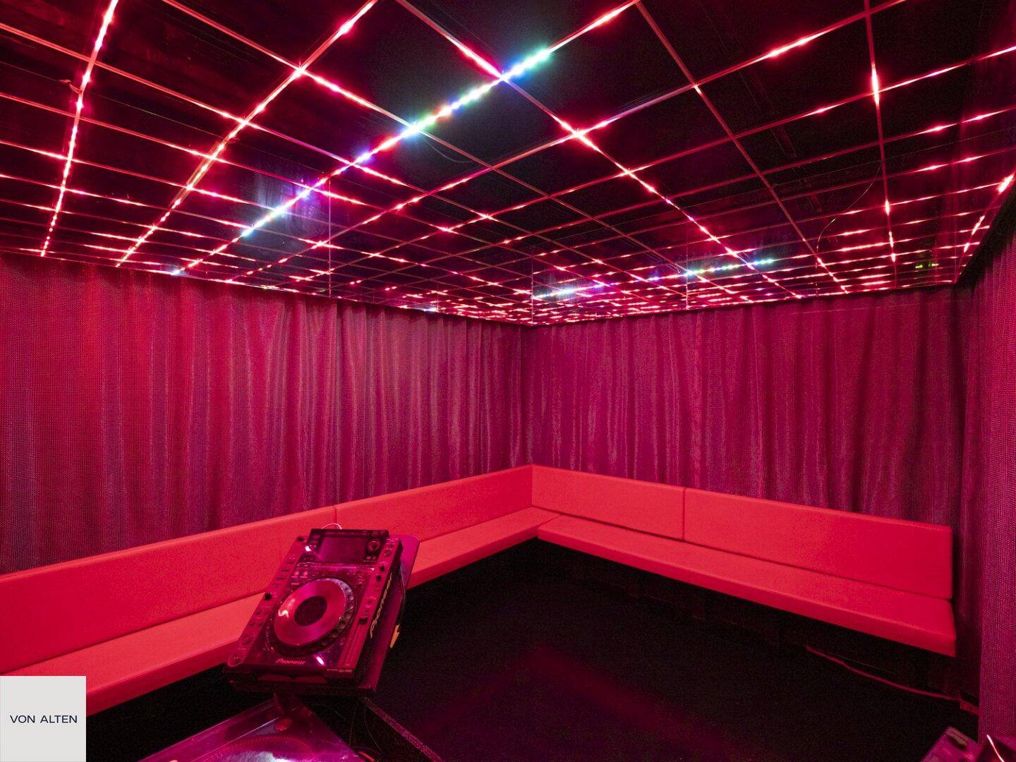 Raum mit roter Beleuchtung, gepolsterten Sitzbänken und DJ-Equipment, ideal für Veranstaltungen.