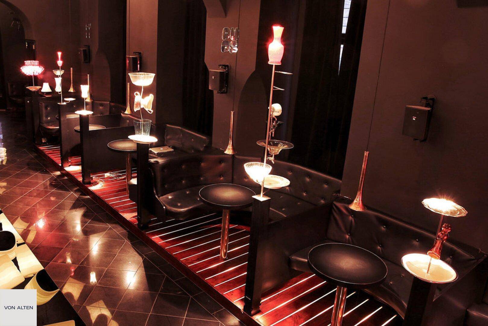 Gemütlicher Sitzbereich in der Bar mit kleinen Tischen, dunklen Bänken und individuellen Lichtobjekten.