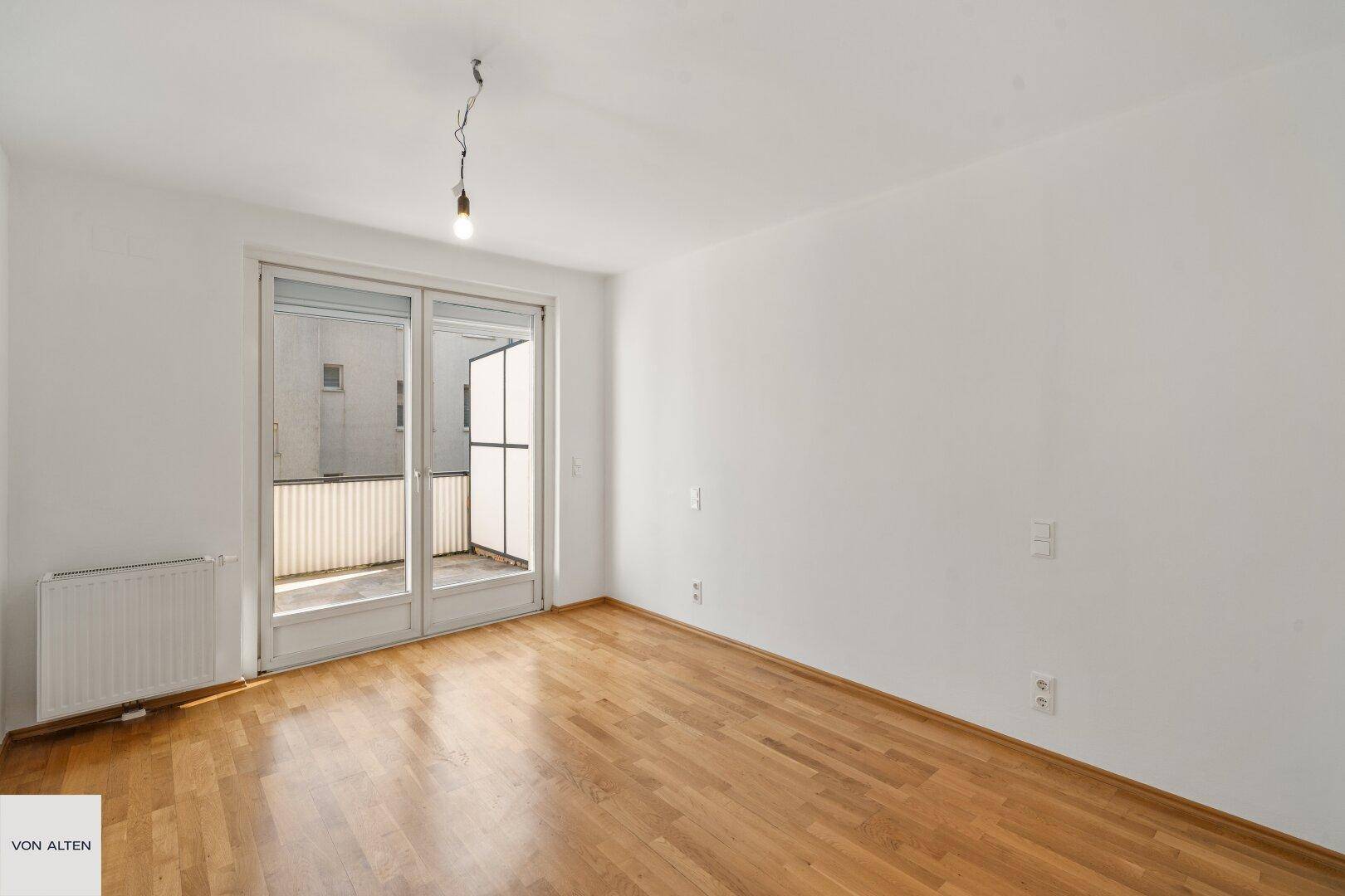 Heller Raum mit Holzboden und Zugang zu einem Balkon oder einer Terrasse.