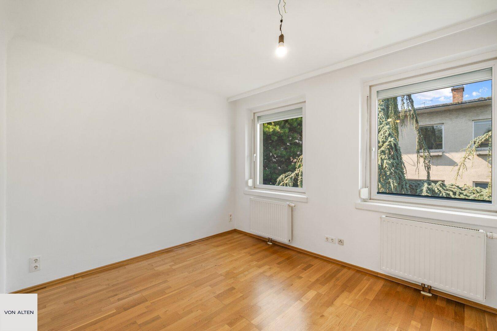 Heller Raum mit Holzboden und zwei Fenstern mit Blick auf Bäume.