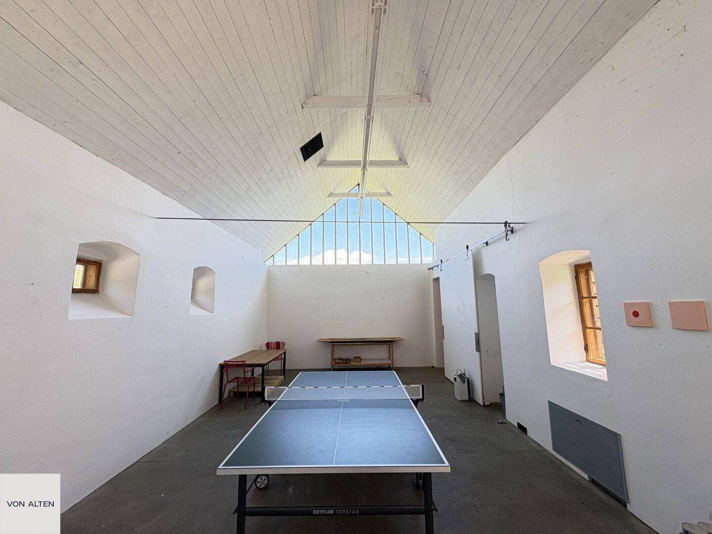 Großer heller Raum mit Tischtennisplatte, hohen Decken und dreieckigem Giebelfenster.