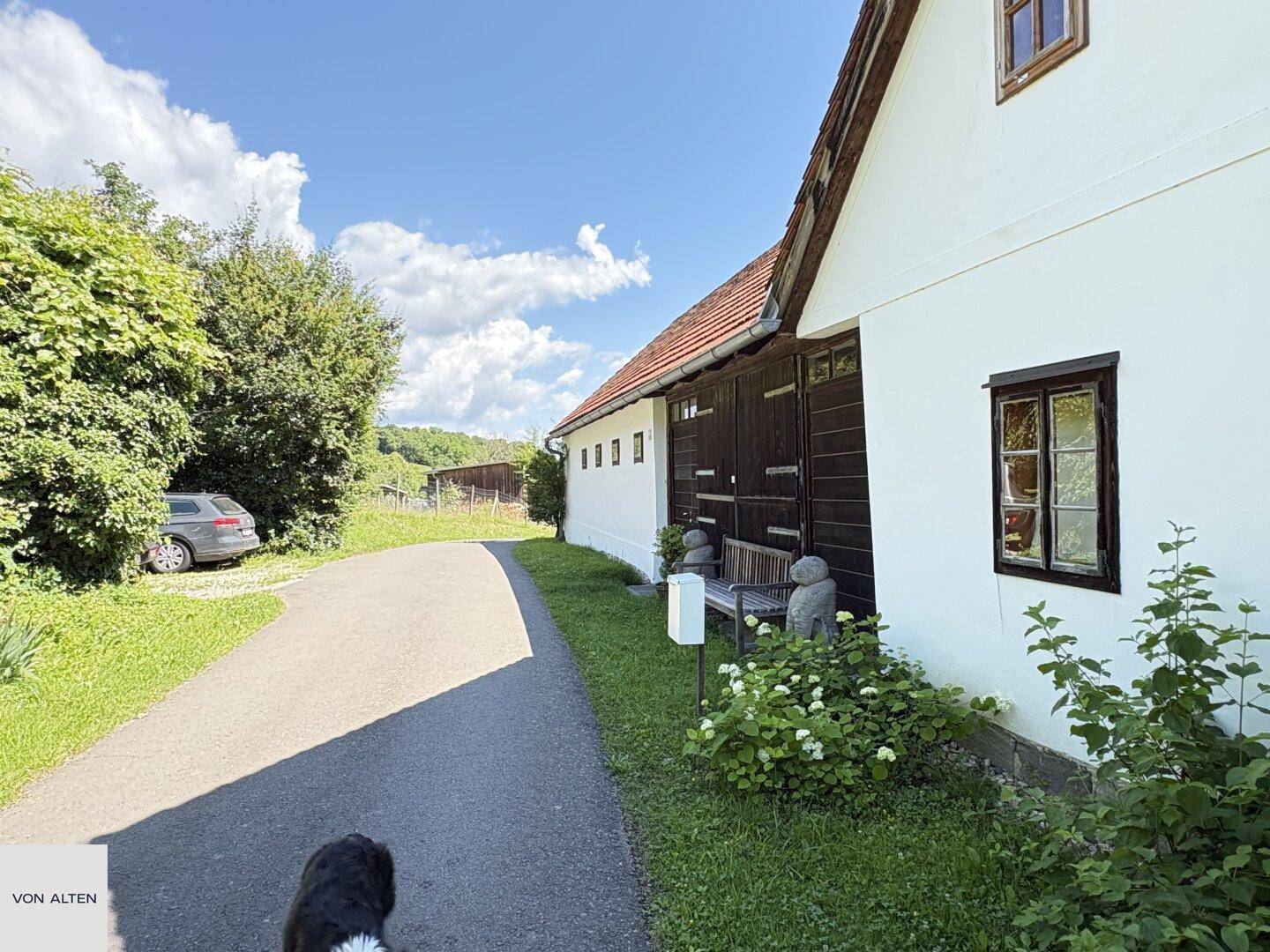Ländlicher Weg führt am weißen Bauernhaus mit Holztüren und grüner Umgebung vorbei.