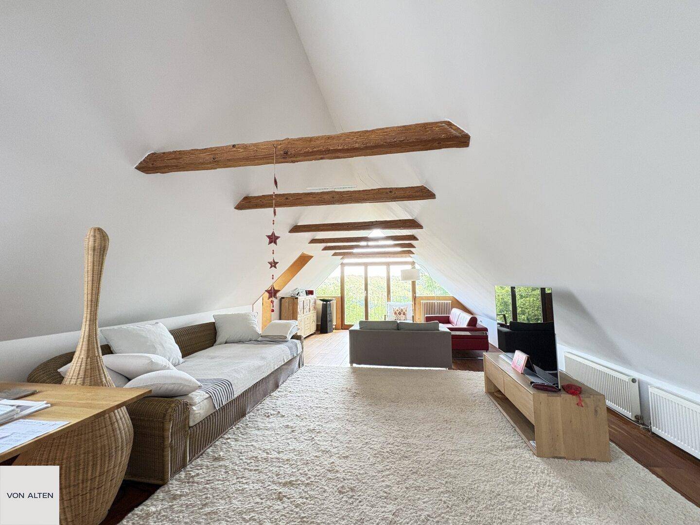 Geräumiges Dachgeschoss-Wohnzimmer mit sichtbaren Holzbalken, Teppichboden und weitem Blick ins Grüne.