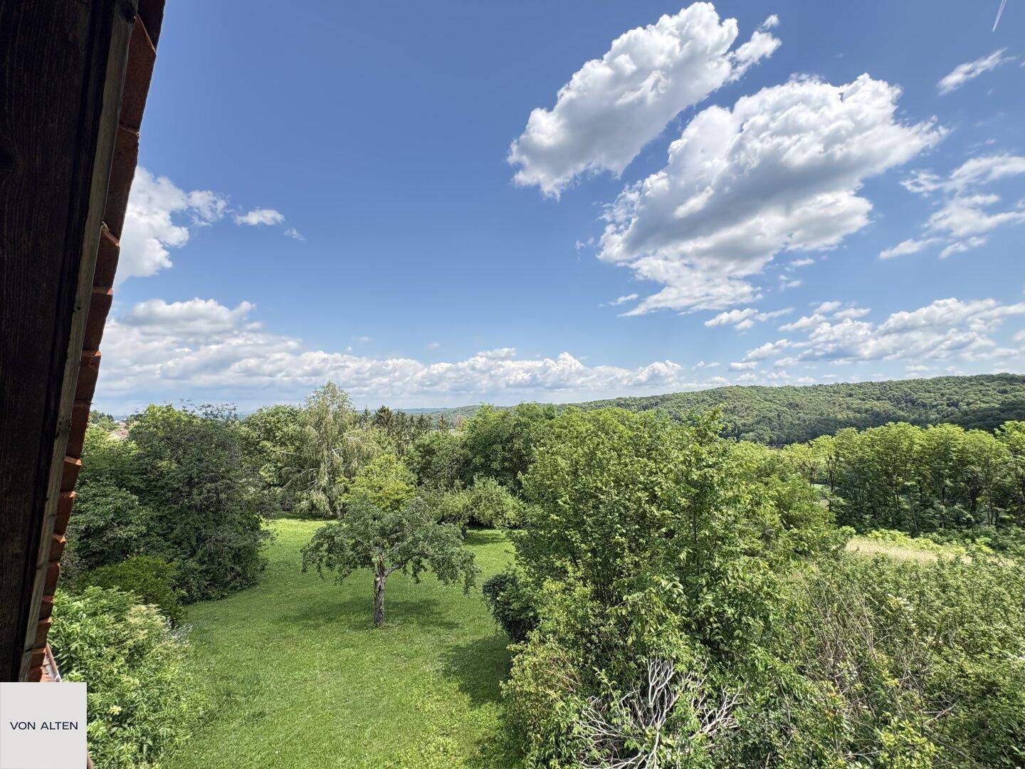 Weitläufiger Blick über eine grüne Landschaft mit Bäumen und Wiesen unter einem sonnigen Himmel.
