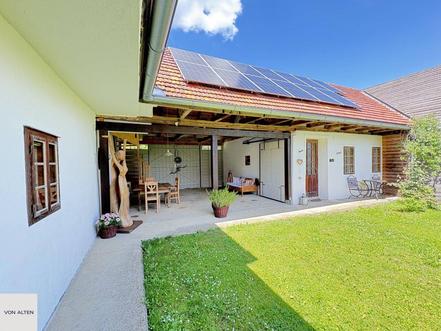 Außenbereich des Hauses mit Rasenfläche, überdachtem Sitzplatz und Solarmodulen auf dem Dach.