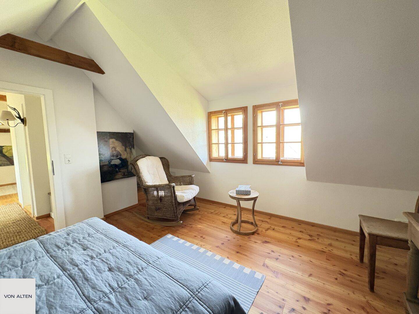Helles Schlafzimmer im Dachgeschoss mit Holzboden, Schaukelstuhl und zwei Fenstern für Tageslicht.