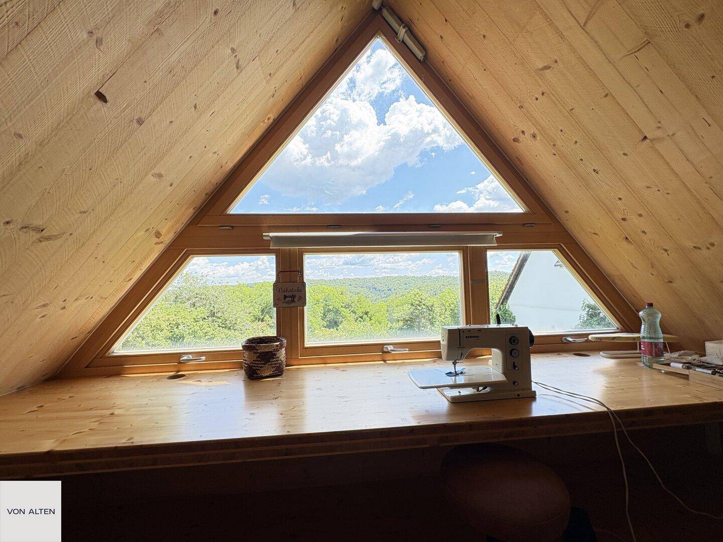 Helle Dachbodenwerkstatt mit Holztisch, Nähmaschine und dreieckigem Fenster mit Weitblick.