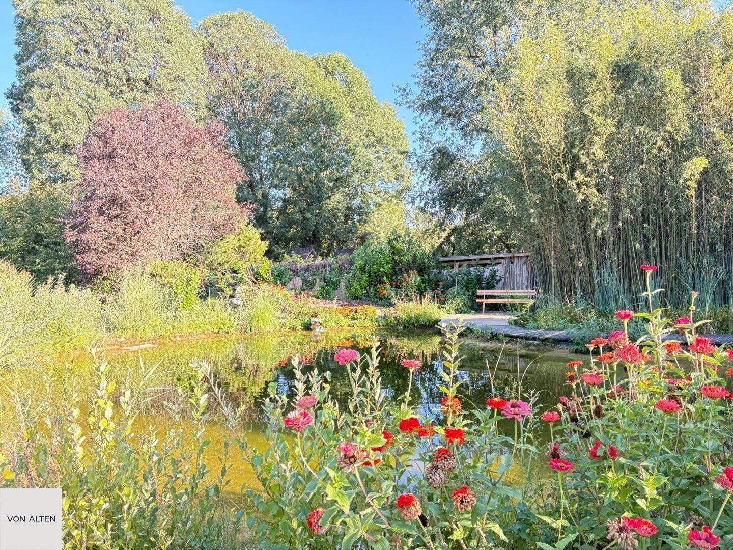 Üppiger Garten mit einem Teich, bunten Blumen und einer Holzbank, umgeben von hohen Bäumen und Bambus.