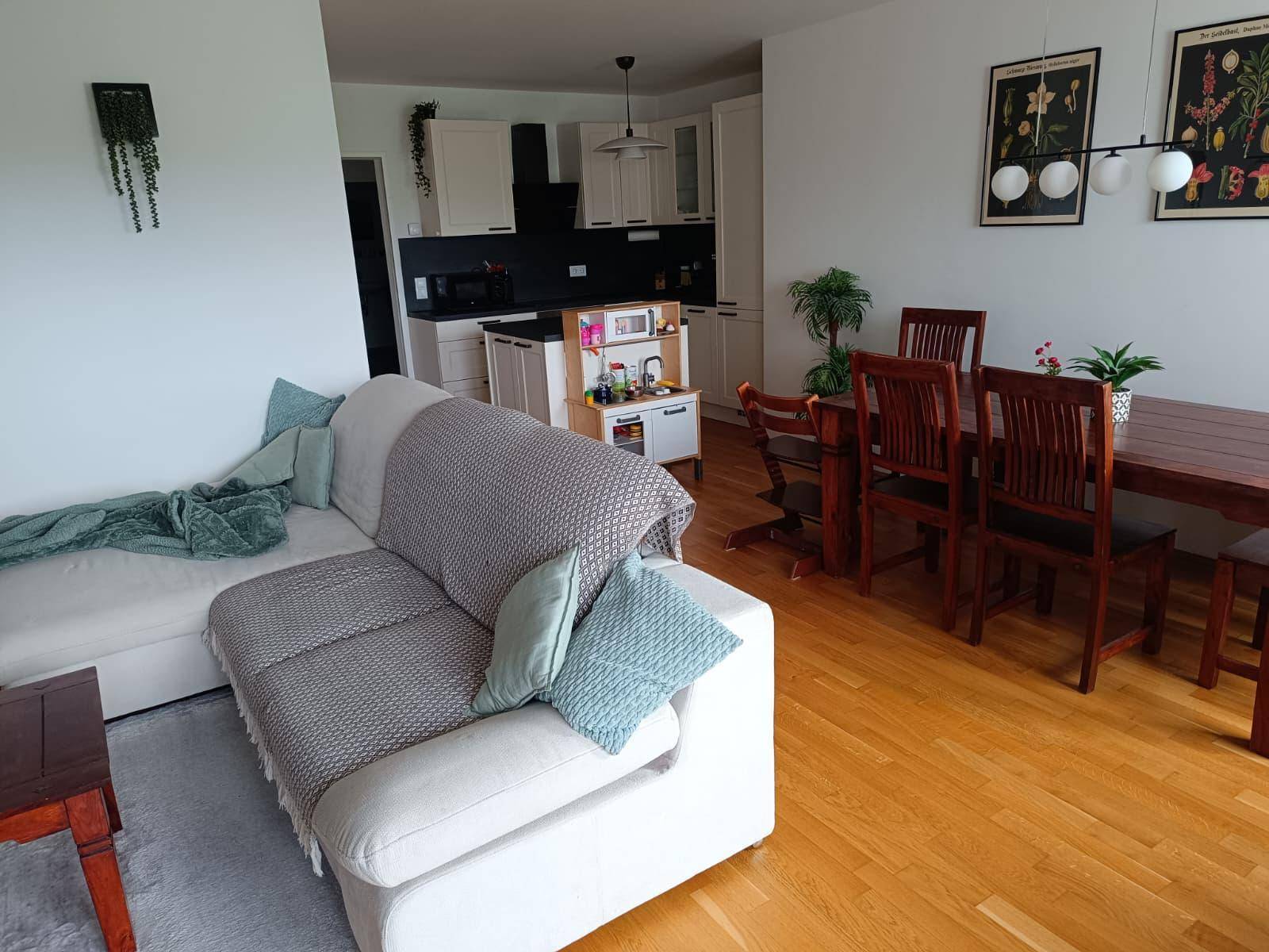 Offener Wohn- und Essbereich mit hellem Sofa, Holztisch und integrierter Küche im Hintergrund.