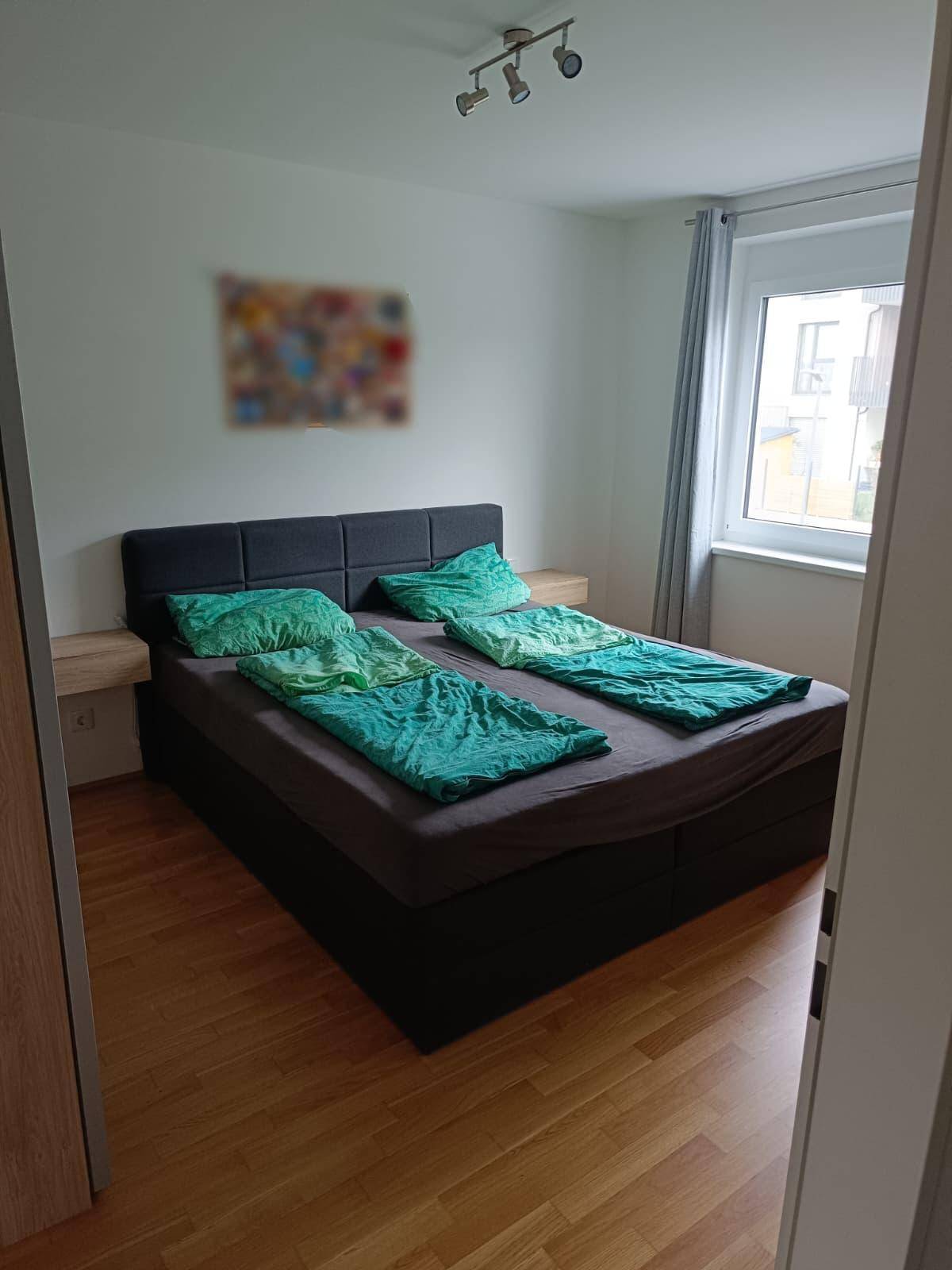 Schlafzimmer mit Doppelbett, Holzboden und einem Fenster, das viel Tageslicht hereinlässt.