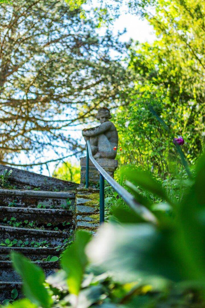 Alte Steintreppe mit Moosbewuchs und einer Statue, umgeben von üppigem Grün im Gartenbereich.