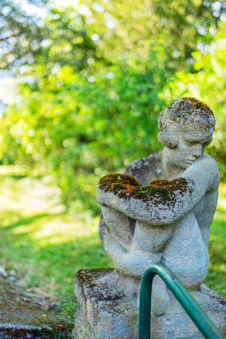 Nahaufnahme einer moosbewachsenen Steinstatue im Garten, umgeben von hellem Grün und Sonnenlicht.