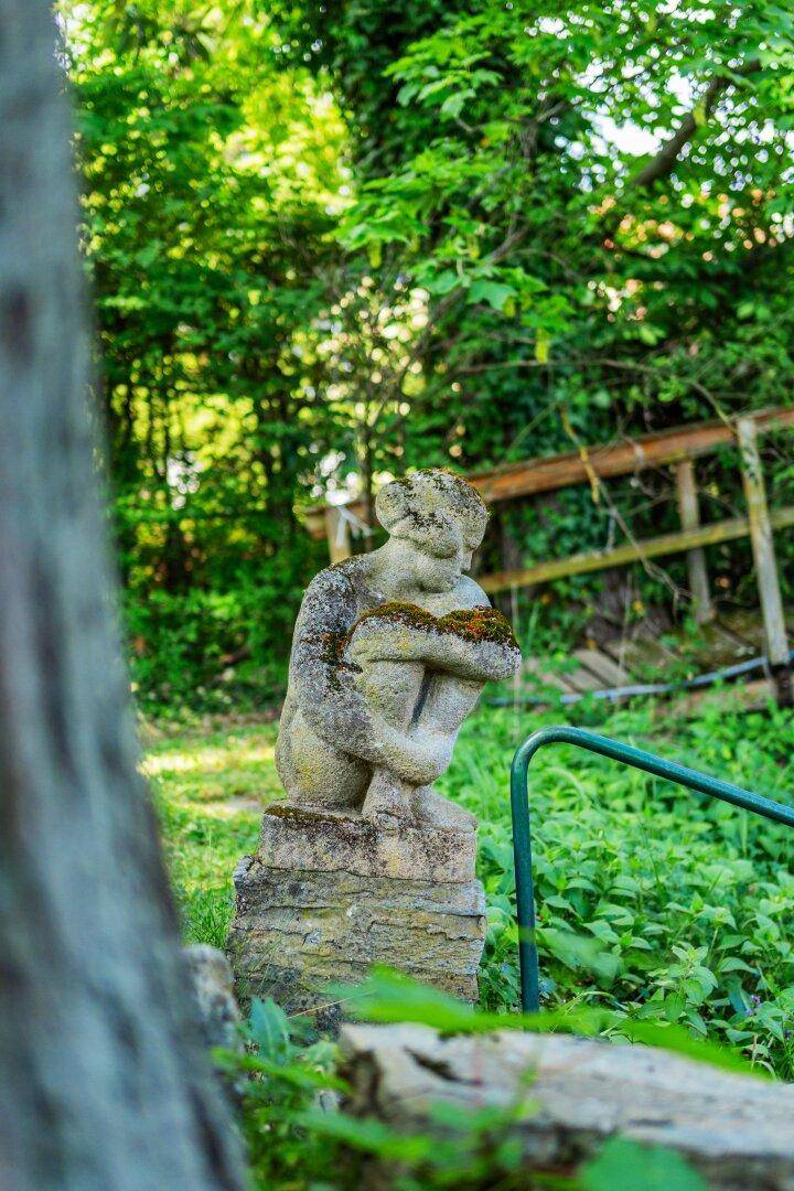 Moosbewachsene Steinstatue in einem verwilderten Gartenbereich mit viel Grün und Bäumen.