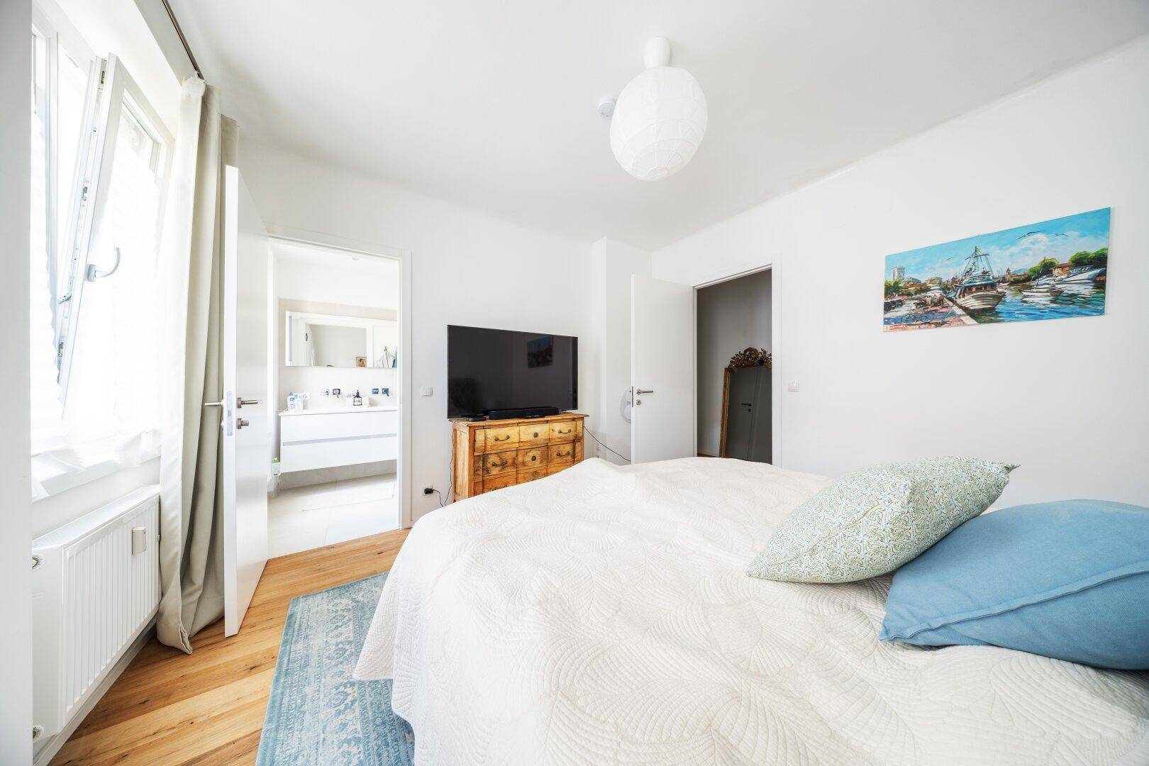 Schlafzimmer mit Zugang zum Badezimmer, ausgestattet mit einem Fernseher und Holzboden.