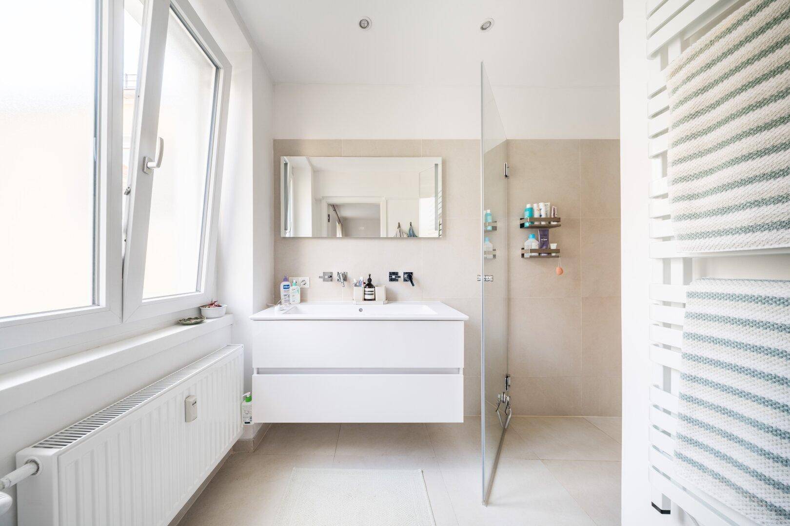 Modernes Badezimmer mit Fenster, begehbarer Dusche, weißem Waschtisch und beheiztem Handtuchhalter.