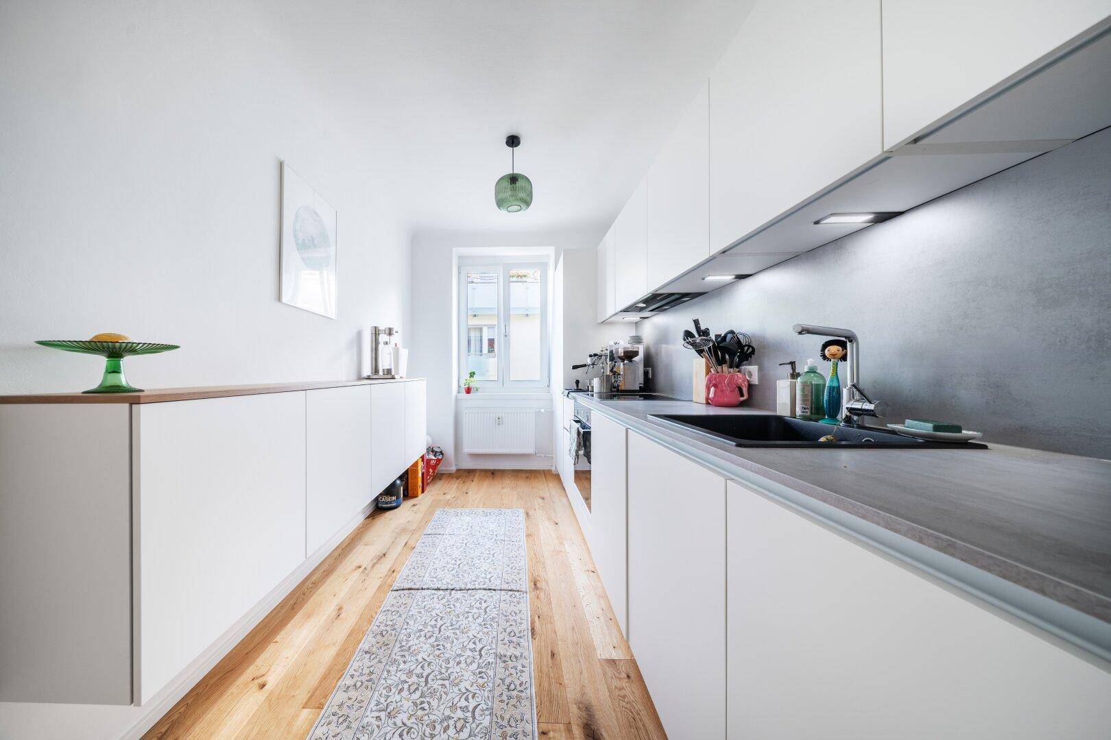 Moderne, schmale Küche mit weißen Schränken, Holzboden und Fenster.