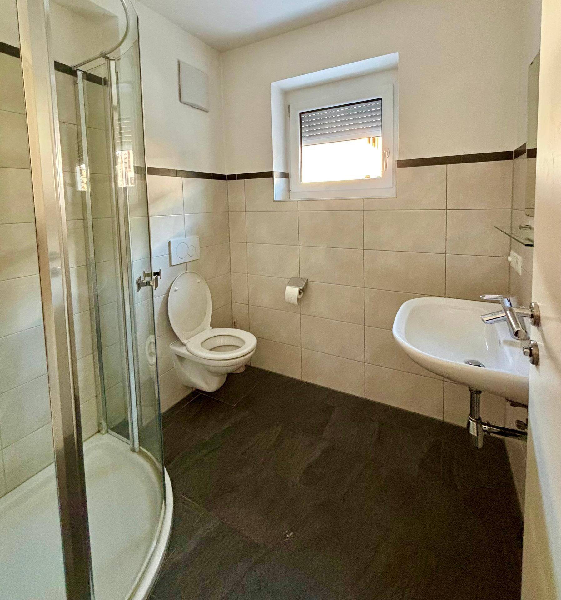 Kleines Badezimmer mit Dusche, Toilette, Waschbecken und Fenster.