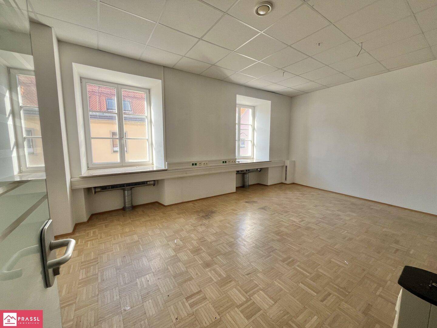 Leerer Büroraum mit Parkettboden, zwei Fenstern und vorbereiteten Anschlüssen an der Wand.
