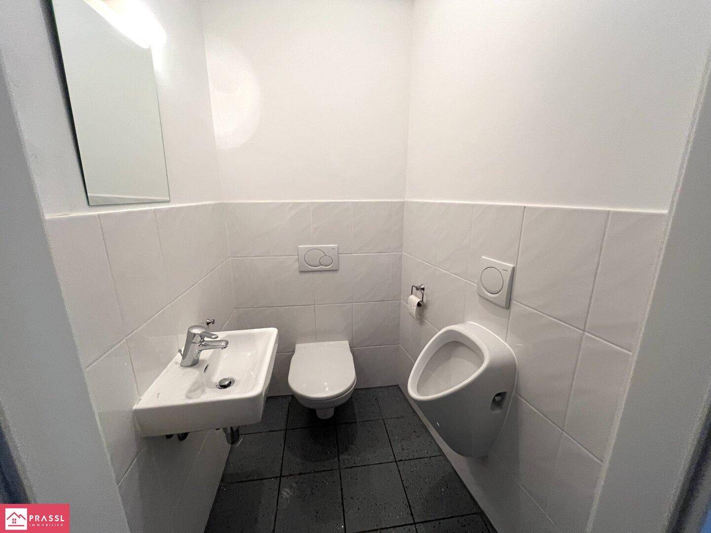 Kleiner Toilettenraum mit weißen Fliesen, Waschbecken, WC und Urinal.