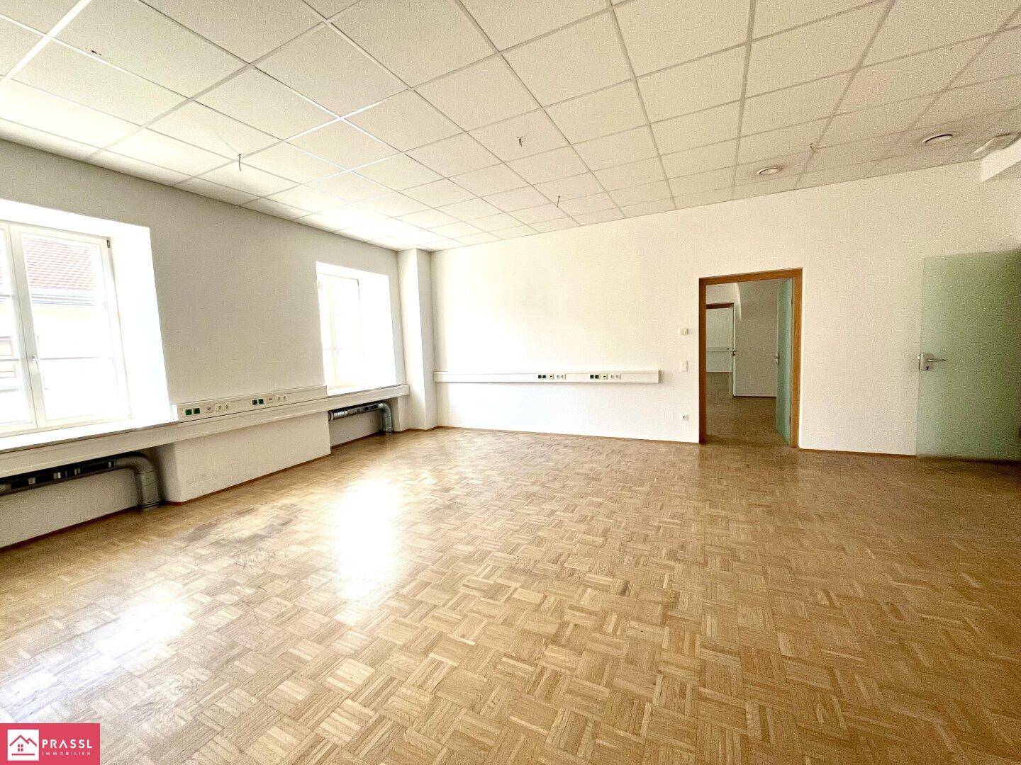 Geräumiger Büroraum mit Parkettboden, großen Fenstern und modularen Deckenplatten.