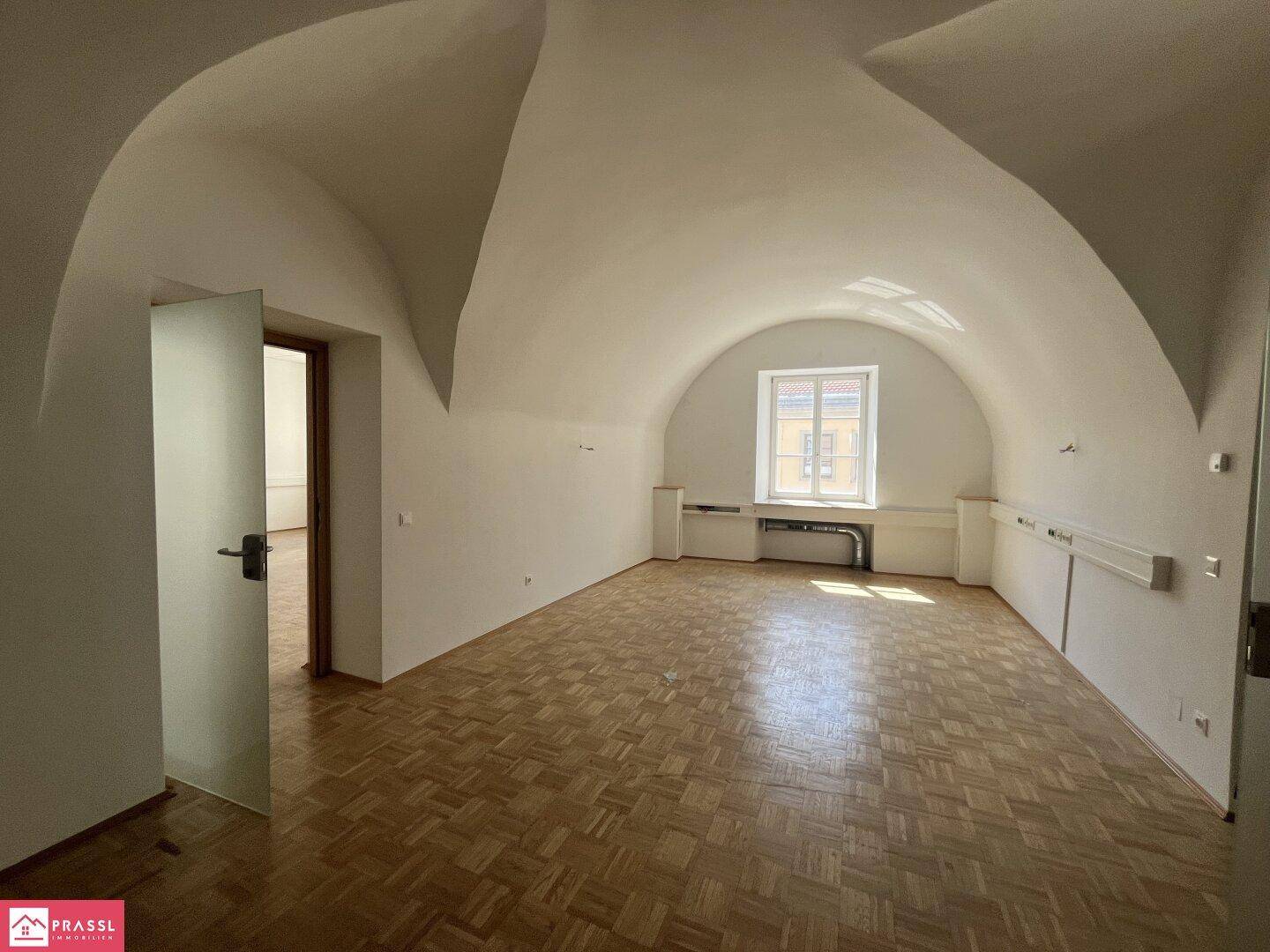 Heller Raum mit beeindruckender Gewölbedecke, Parkettboden und einem Fenster mit Blick nach draußen.