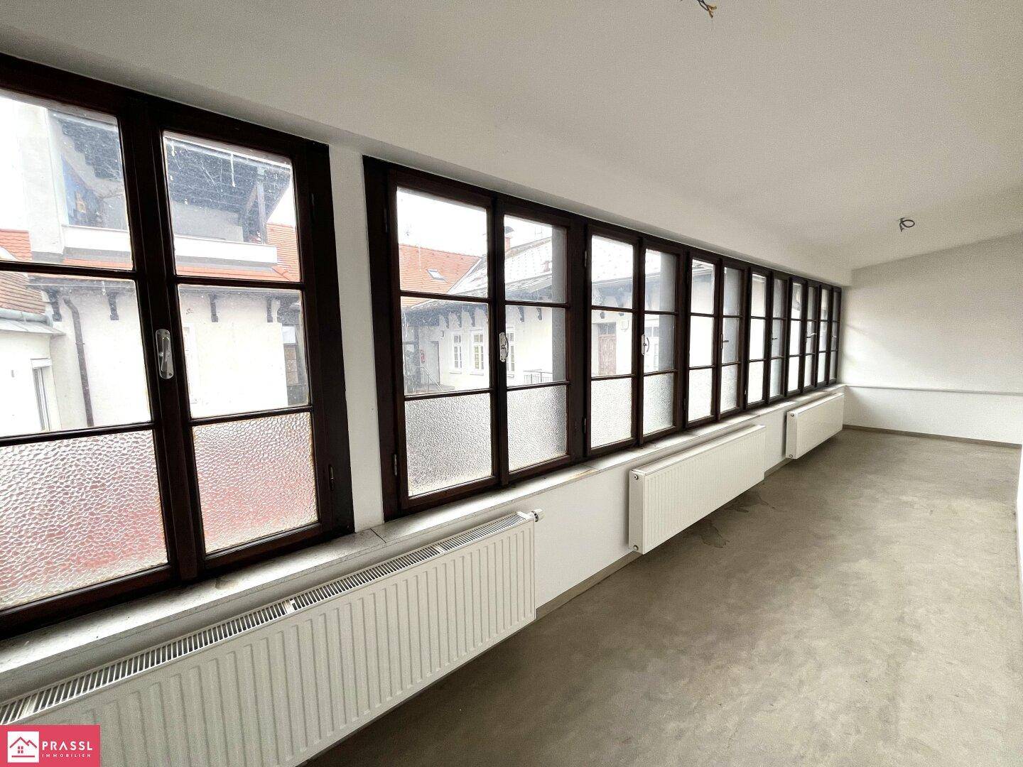 Langer Raum mit vielen Fenstern, die viel Tageslicht hereinlassen, und Heizkörpern darunter.