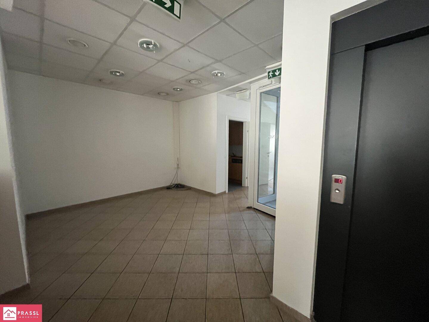 Leerer Gewerberaum mit Fliesenboden, einem modernen Aufzug und Zugang zu einer kleinen Küchenzeile.