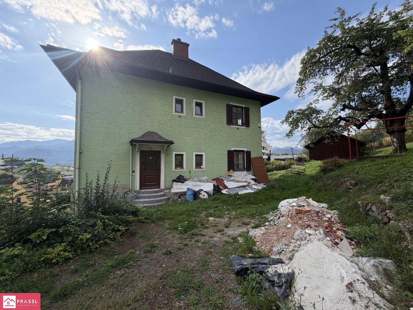 Grünes Haus mit Baustellenresten im Vordergrund, umgeben von Bäumen und Hügeln.