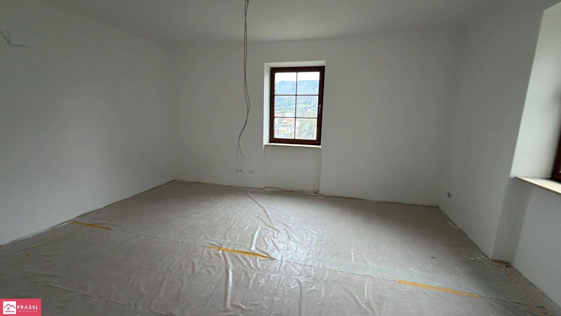 Renovierungsbedürftiger Raum mit einem Fenster, weiß gestrichenen Wänden und abgedecktem Boden.