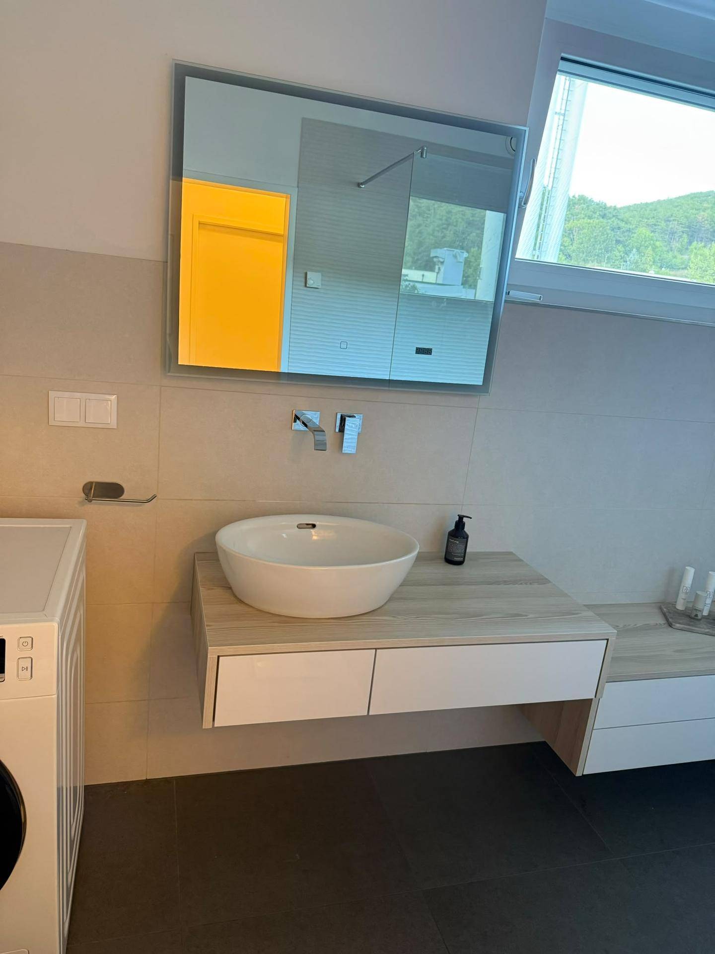 Modernes Badezimmer mit Waschbecken, Spiegel und Fenster, hell gefliest.