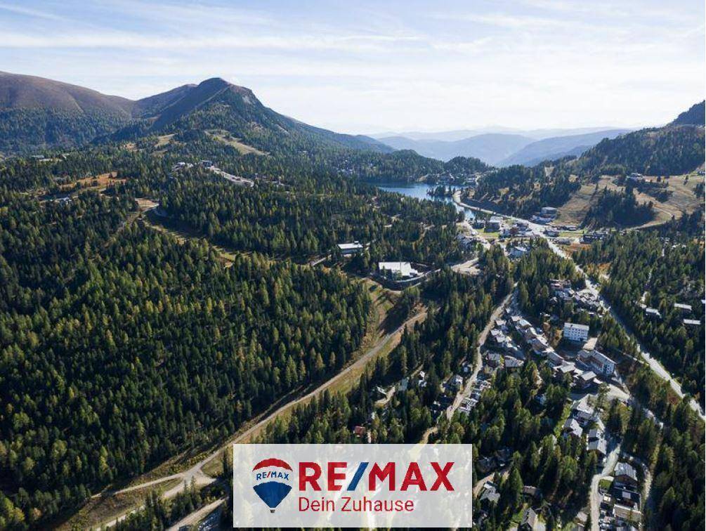 Weitläufige Luftaufnahme einer Berglandschaft mit dichten Wäldern, einem See und Siedlungen unter blauem Himmel.