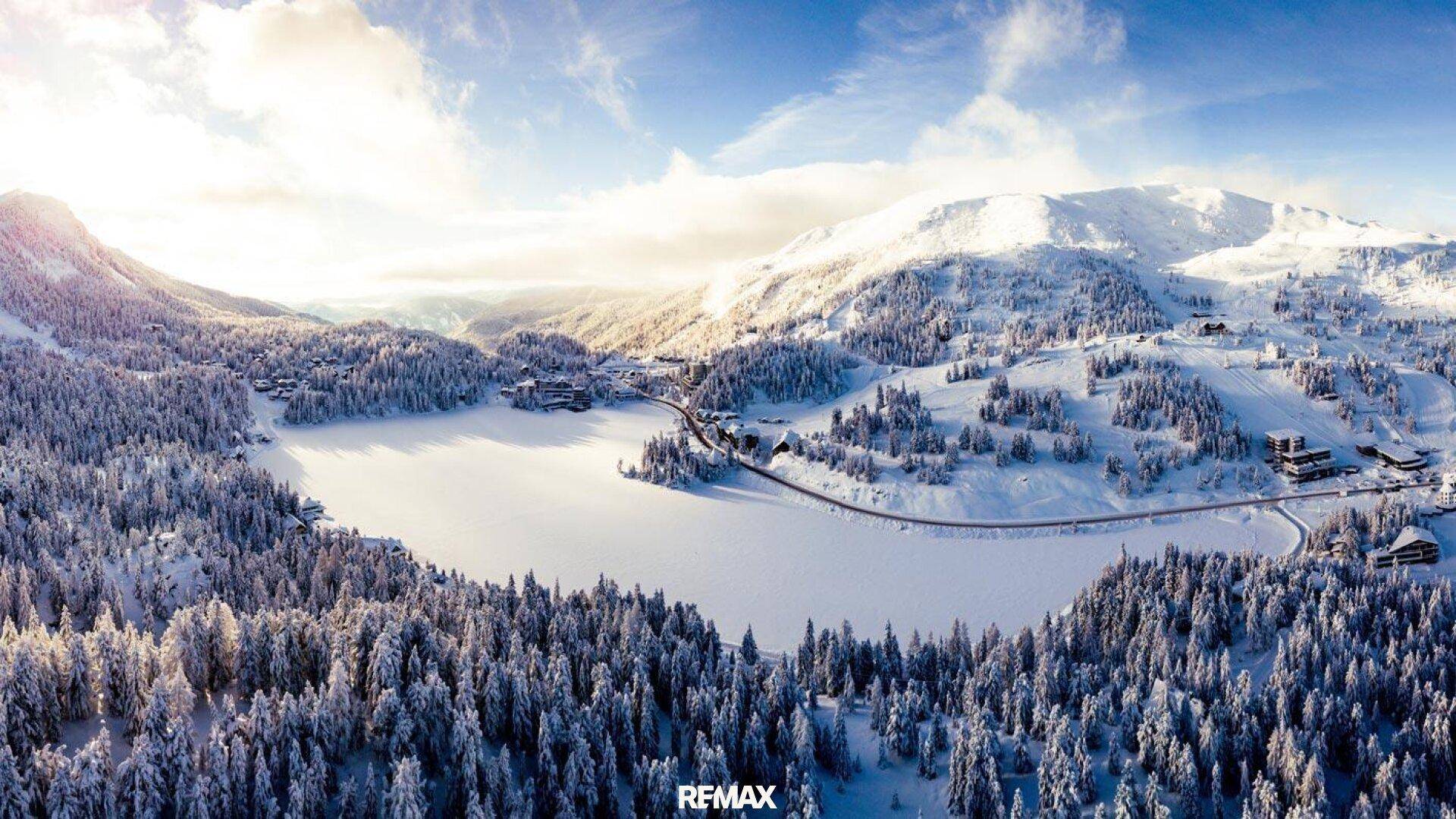 Winterliche Luftaufnahme einer verschneiten Berglandschaft mit zugefrorenem See und Wäldern.