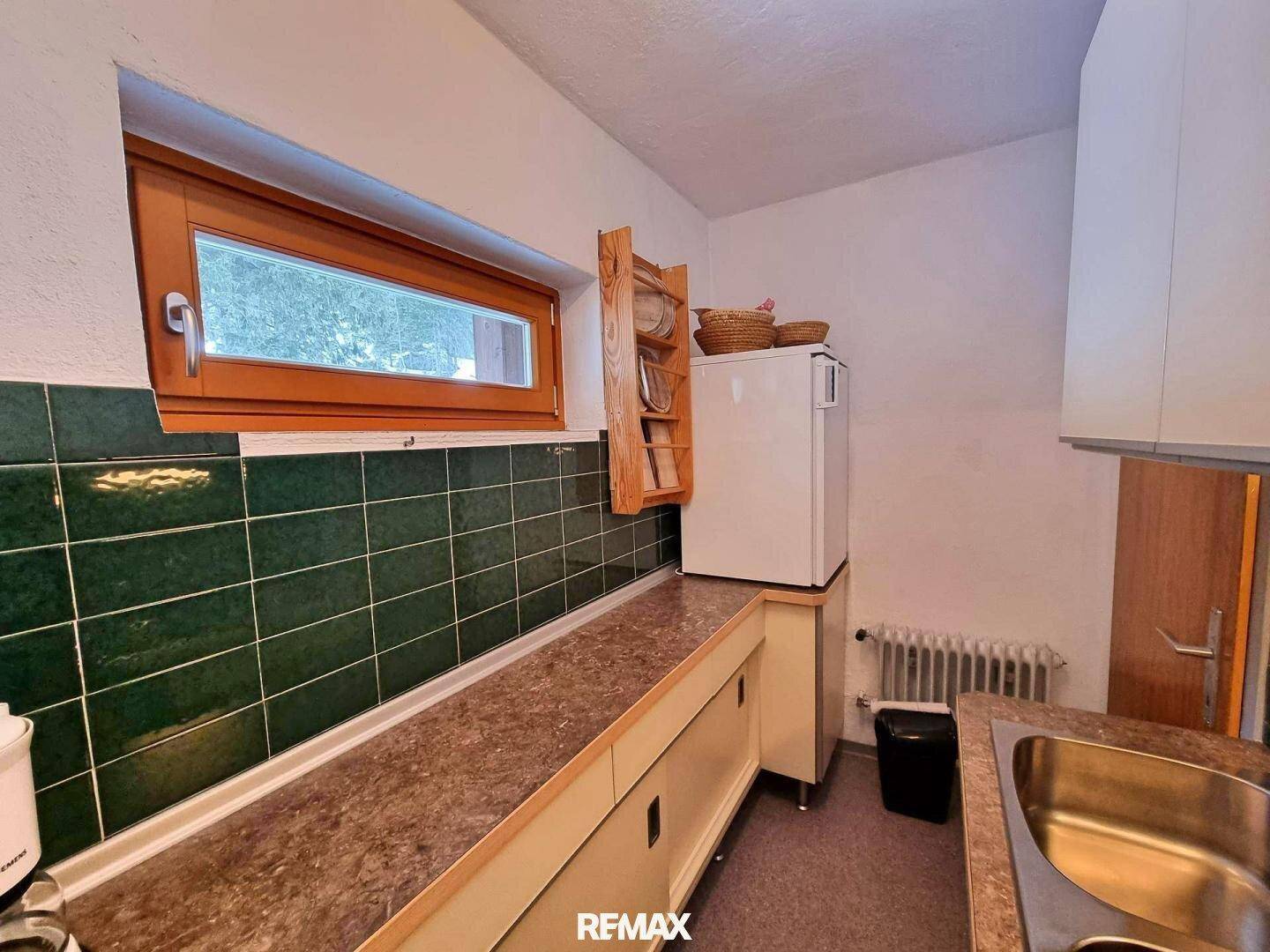 Einfache Küche mit grünen Fliesen, Holzfenster und kleinem Kühlschrank.