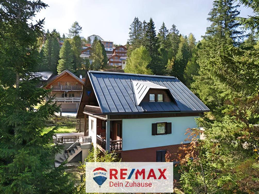 Chalet mit blauem Metalldach und Holzbalkon, eingebettet in eine bewaldete Berglandschaft mit weiteren Gebäuden.
