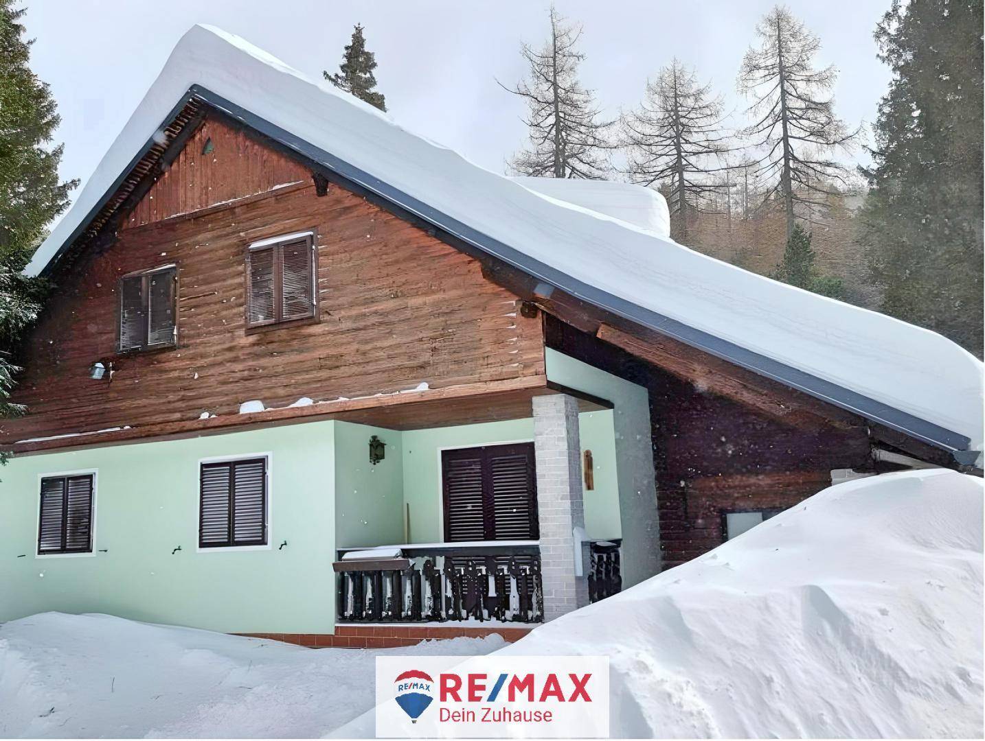 Chalet mit schneebedecktem Dach und Holzbalkon, umgeben von tiefem Winterschnee und Tannenbäumen.