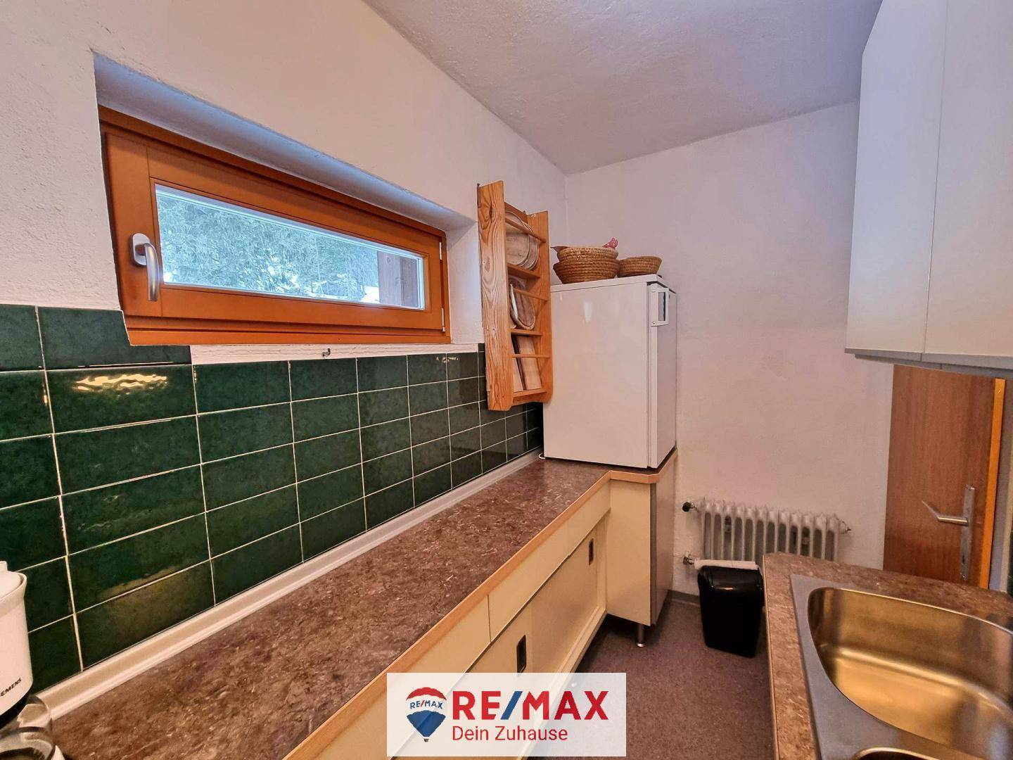 Einfache Küche mit grünen Wandfliesen, Spüle und einem Fenster mit Blick ins Grüne.