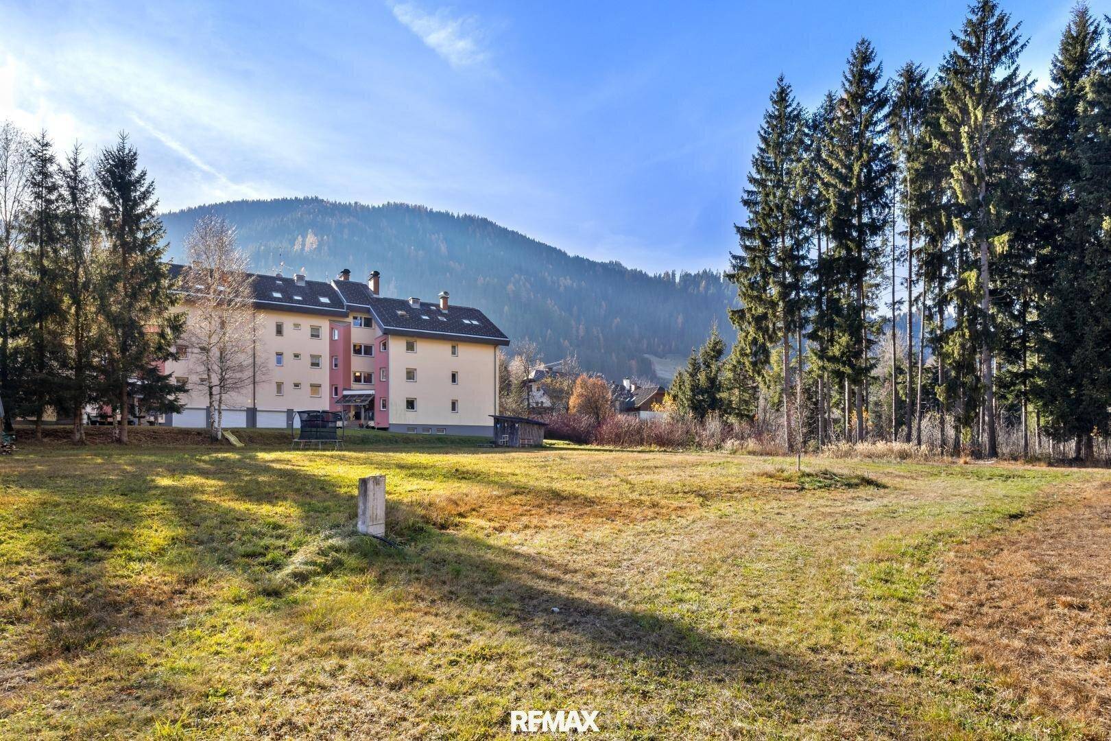 Offenes, grünes Grundstück mit einem Mehrfamilienhaus und hohen Bäumen vor einer Berglandschaft.