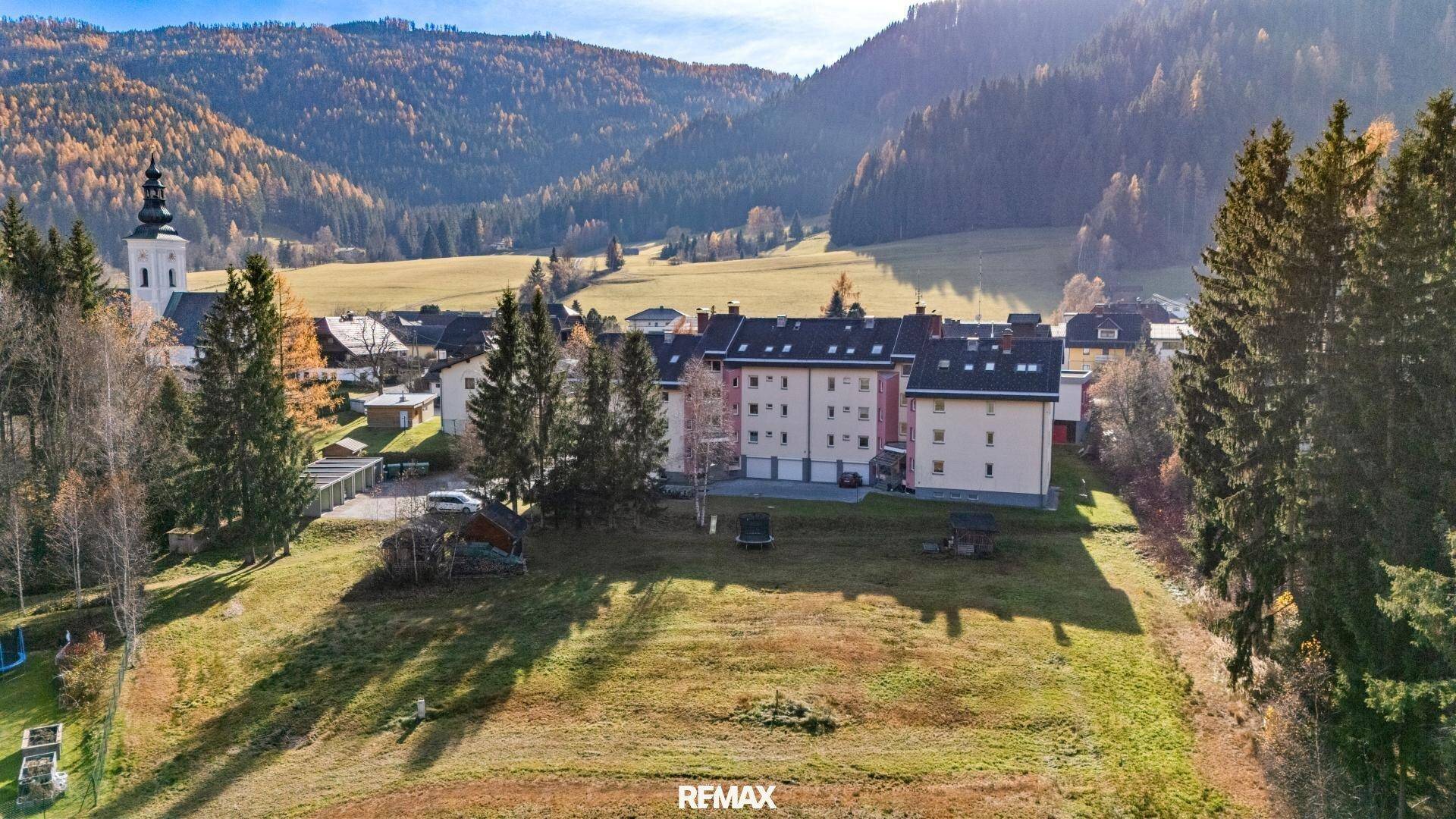 Luftaufnahme des Grundstücks mit angrenzenden Gebäuden, Kirche und bewaldeten Bergen im Hintergrund.