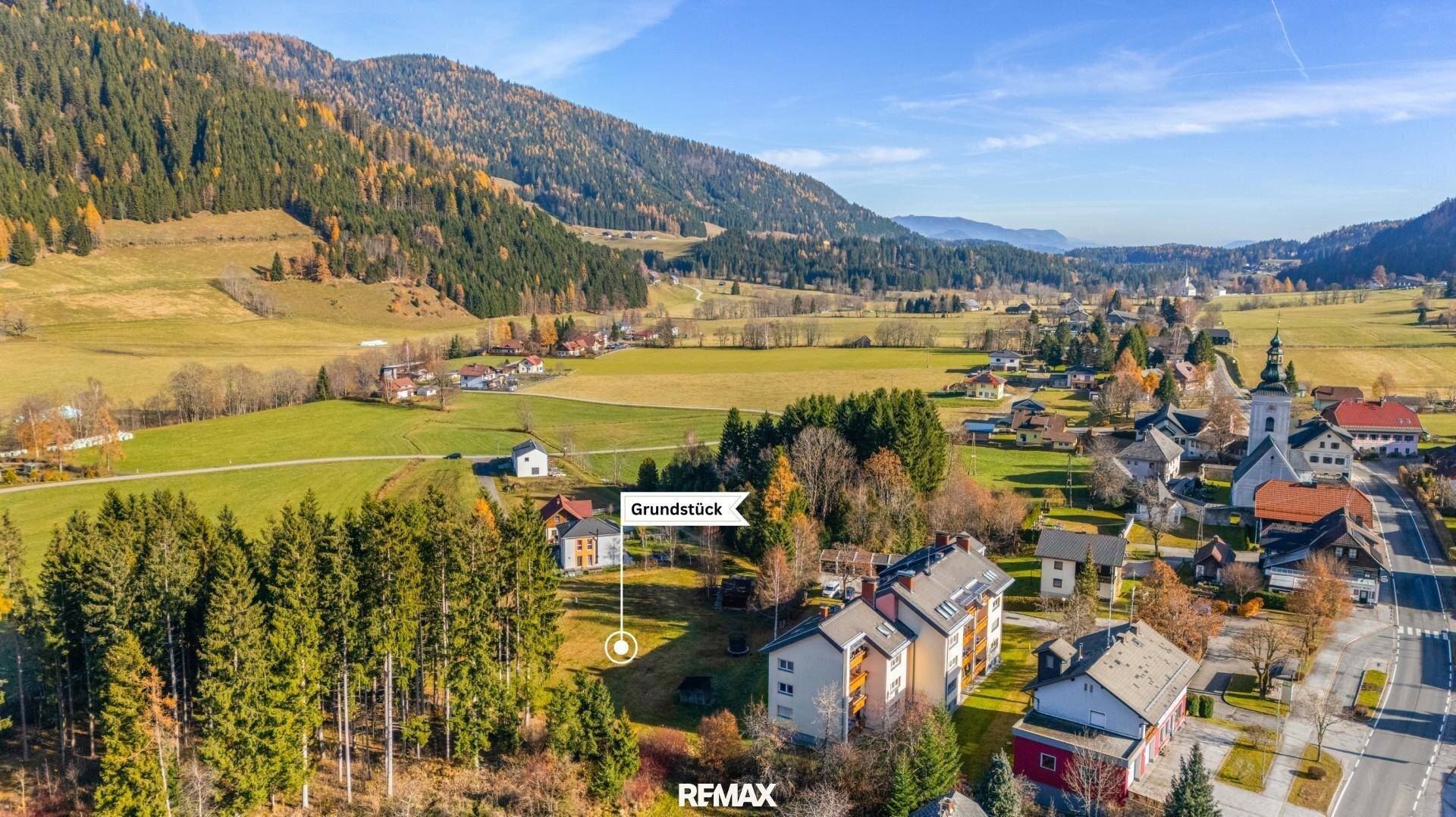 Weitläufige Ansicht eines Bergdorfes mit grünen Hängen und einem markierten Baugrundstück.