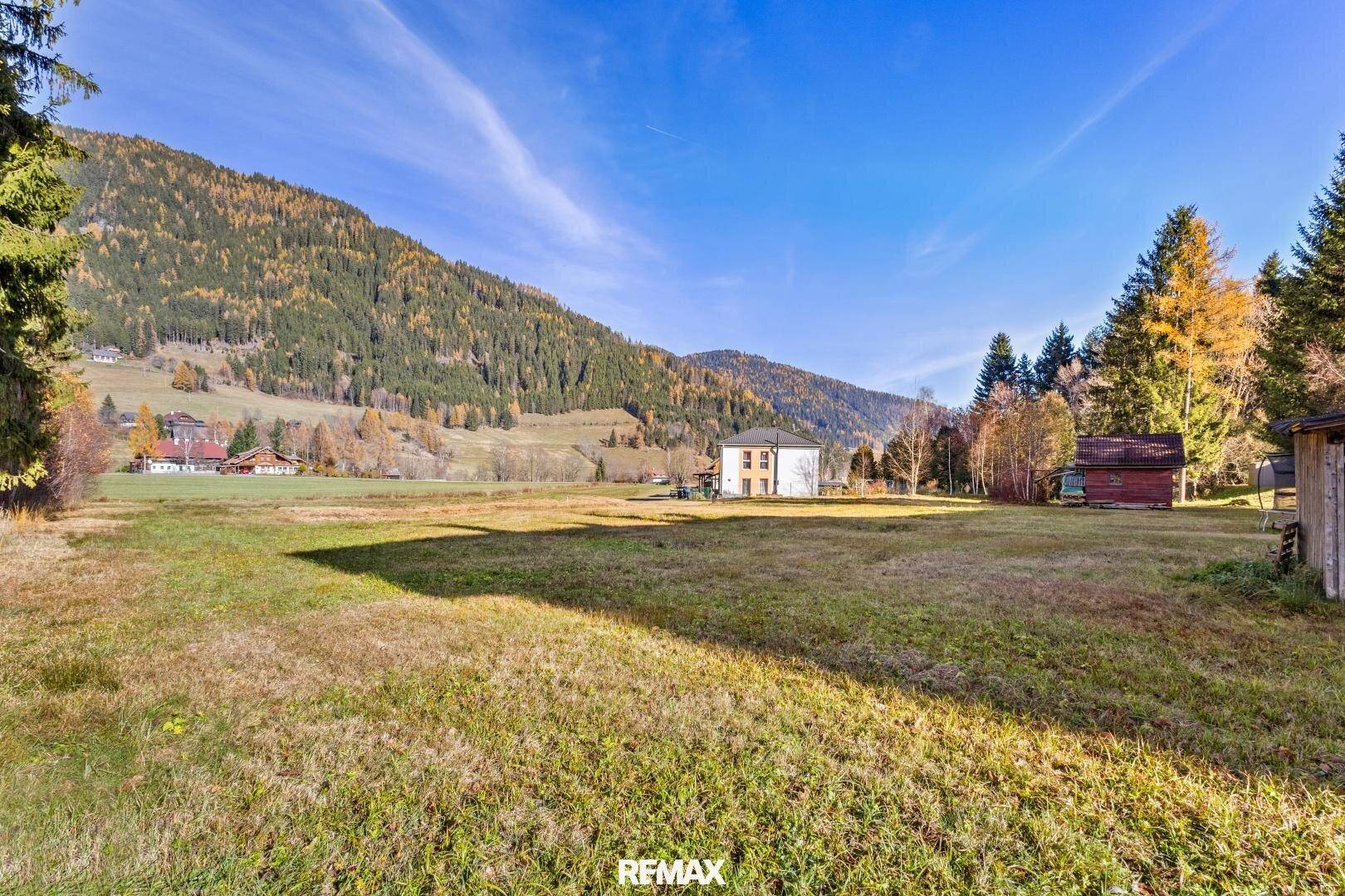 Weitläufiges grünes Grundstück mit einem Haus und hohen Bäumen vor einer Bergkulisse.
