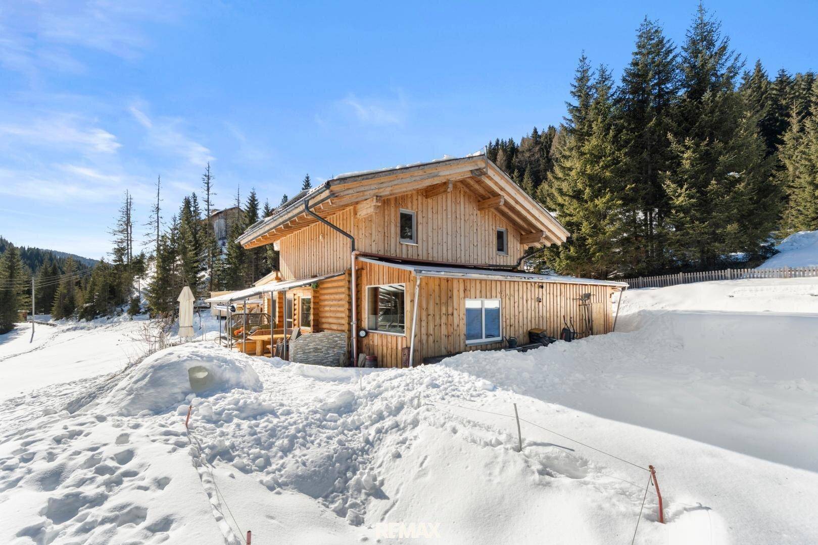 Rustikales Holzchalet in einer tief verschneiten Berglandschaft, umgeben von Nadelbäumen.