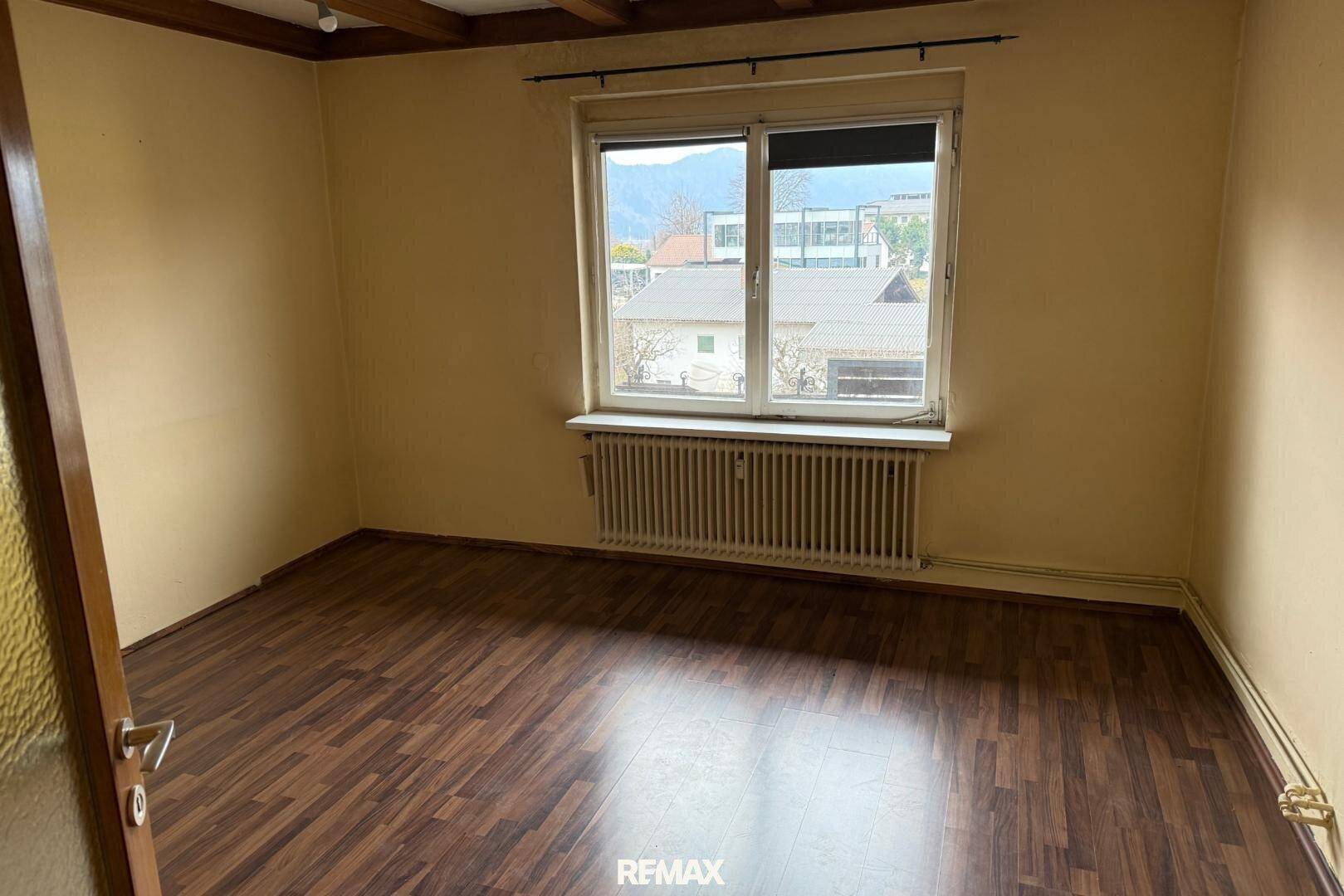 Schlafzimmer mit Laminatboden, Fenster und Heizkörper, das eine Renovierung erfordert.