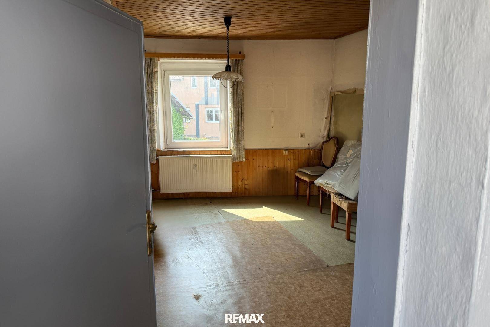 Einfacher Raum mit Holzdecke, Fenster und Heizkörper, der auf eine Renovierung wartet.