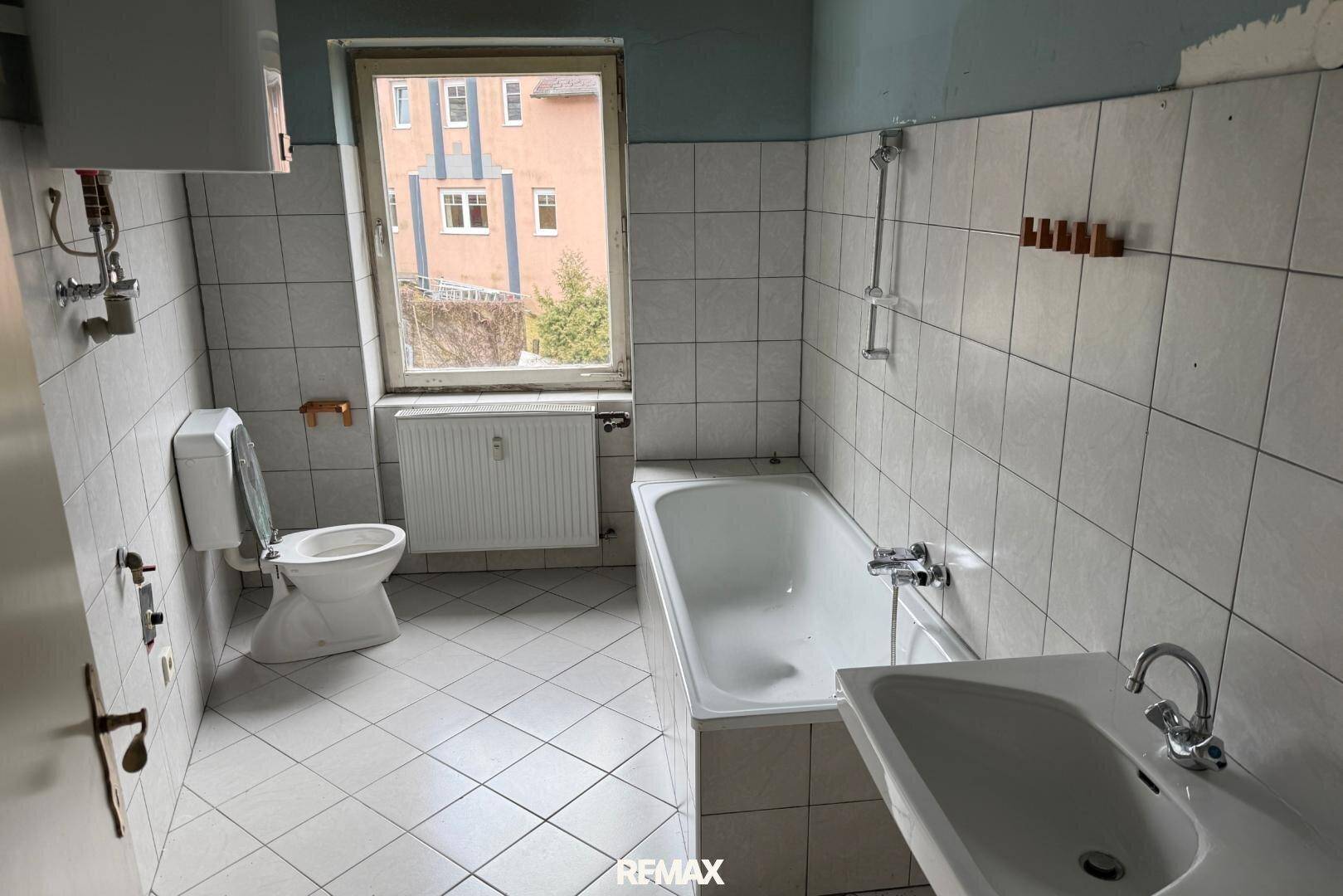 Gefliestes Badezimmer mit Badewanne, Toilette und Fenster, das eine Modernisierung benötigt.