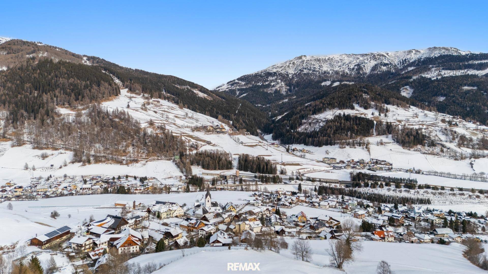 Panoramablick auf ein verschneites Alpendorf, umgeben von Bergen und Wäldern.