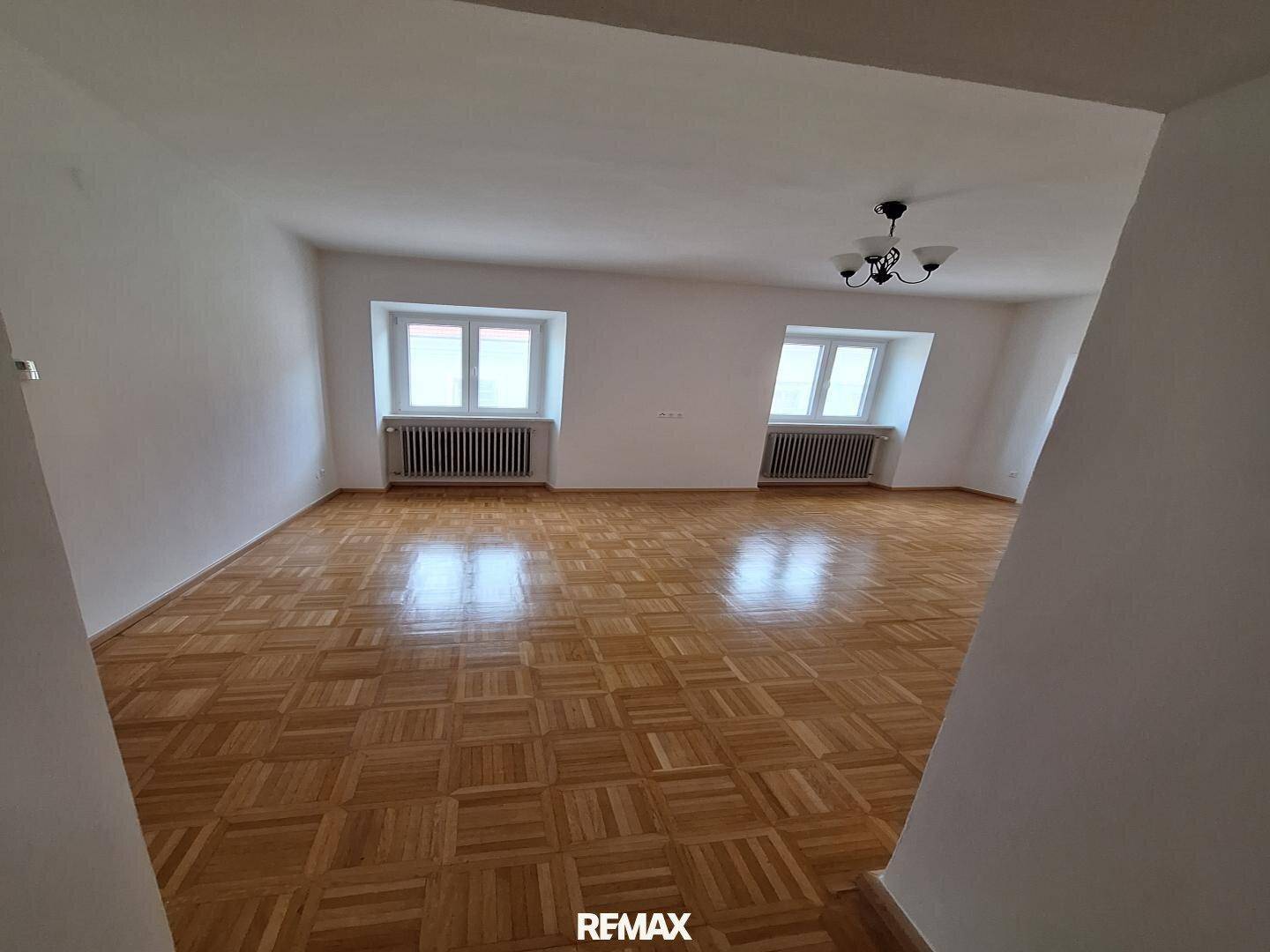 Geräumiges Zimmer mit glänzendem Parkettboden, weißen Wänden und zwei Fenstern mit Heizkörpern.