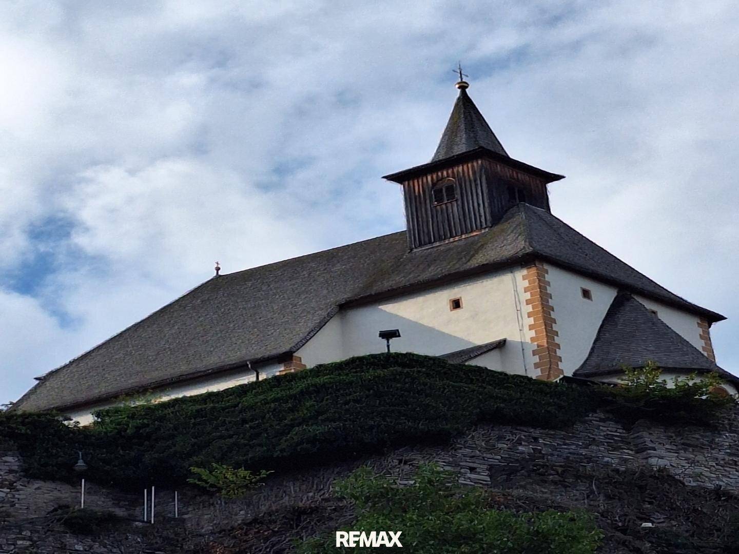 Historische Kirche auf einem Hügel unter bewölktem Himmel, umgeben von grüner Vegetation.