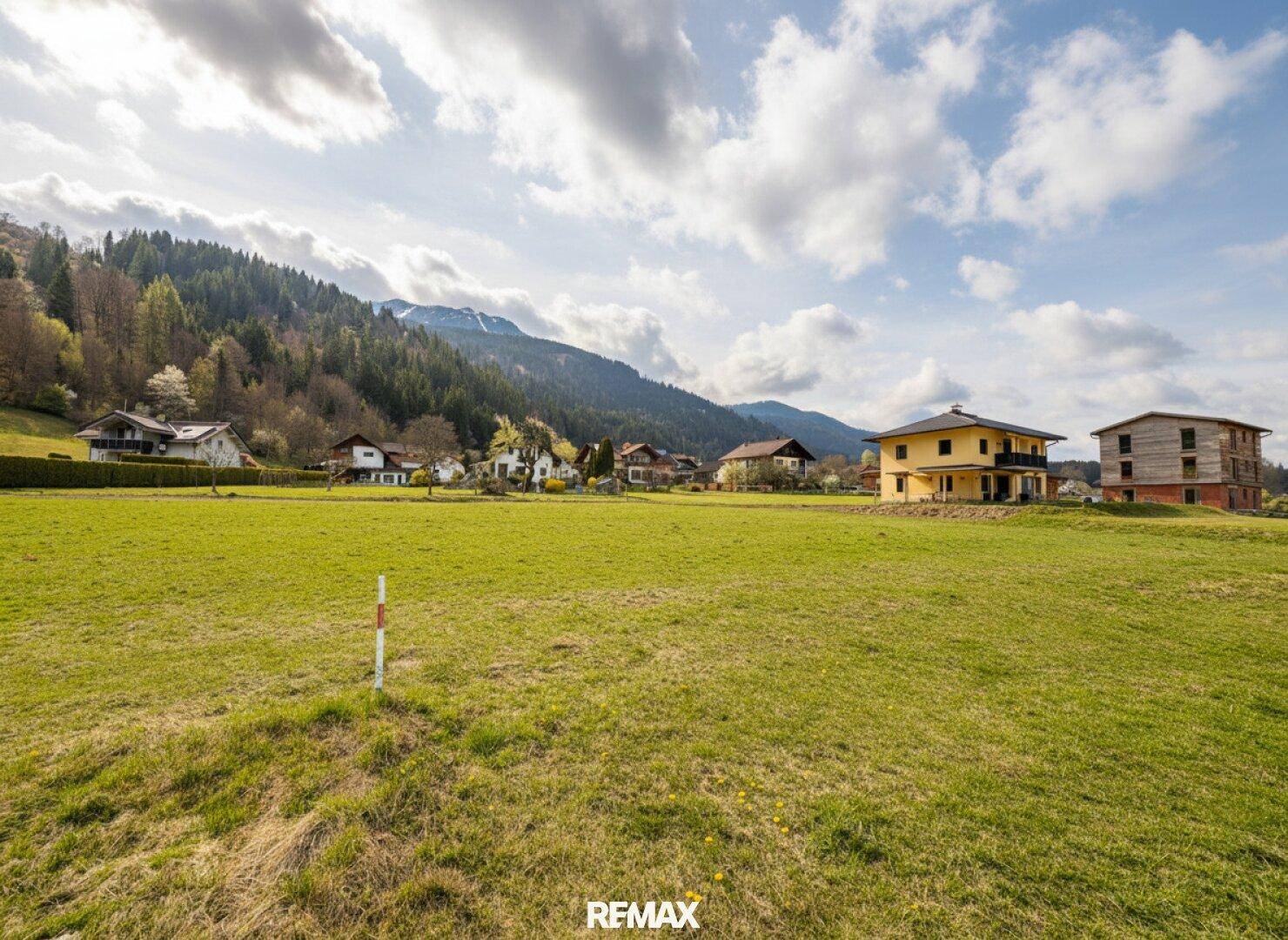 Blick über grüne Wiesen auf eine Ansammlung von Häusern am Fuße bewaldeter Berge unter blauem Himmel.